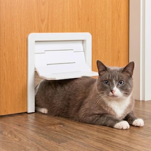 Könnte beinhalten: Eine weiße Haustierklappe aus Kunststoff, die in einen hölzernen Türrahmen eingebaut ist. Eine grau-weiße Katze liegt vor der Haustierklappe, deren Klappe teilweise geöffnet ist. Die Klappe hat einen rechteckigen Rahmen und einen Scharniermechanismus.