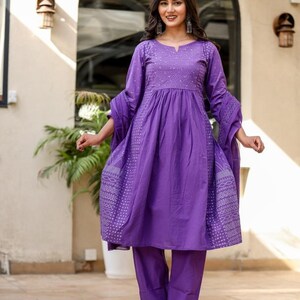 May include: A vibrant purple dress and trouser set. The dress has long sleeves and white embroidered patterns. The outfit is completed with matching trousers and silver sandals. The dress and trousers are a set.