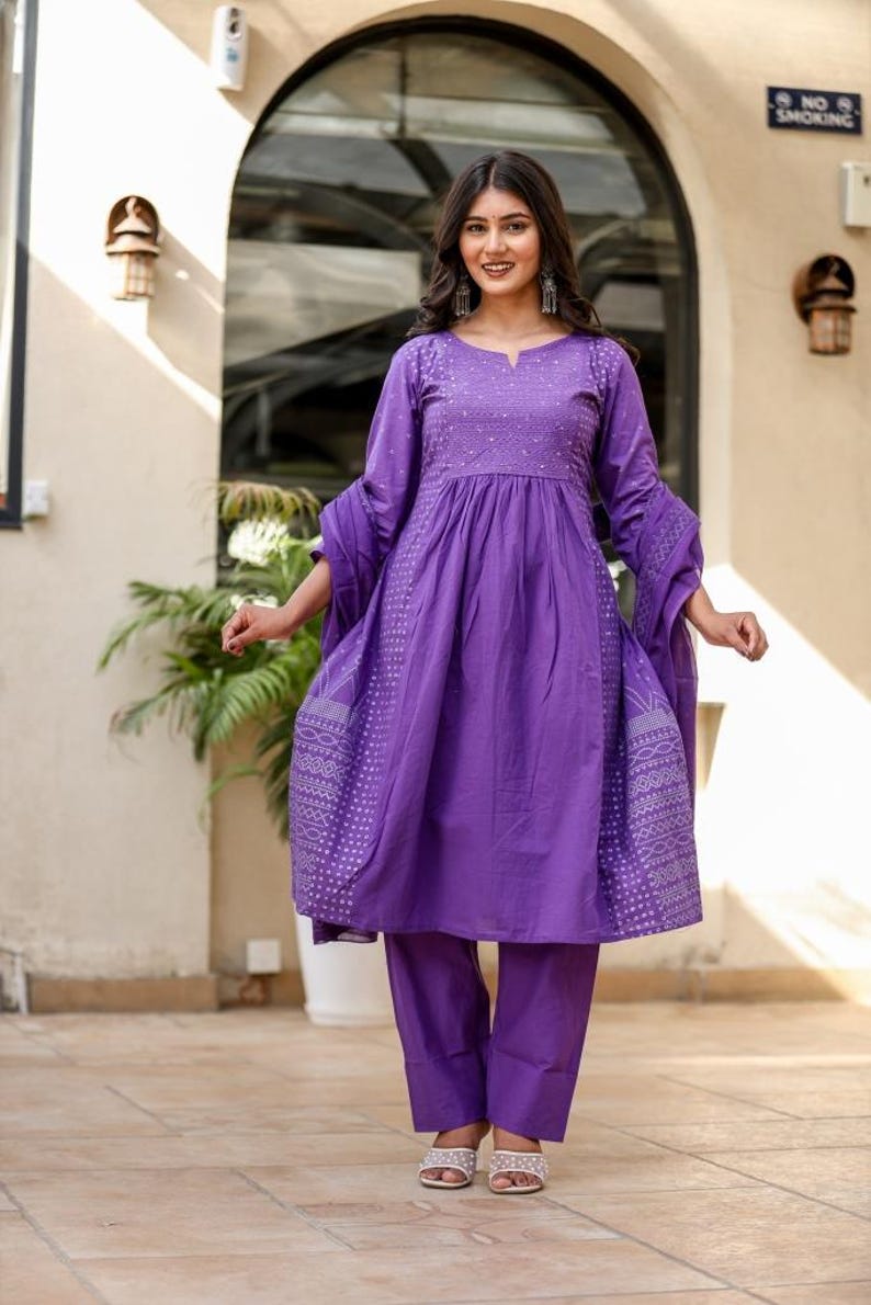 May include: A vibrant purple dress and trouser set. The dress has long sleeves and white embroidered patterns. The outfit is completed with matching trousers and silver sandals. The dress and trousers are a set.
