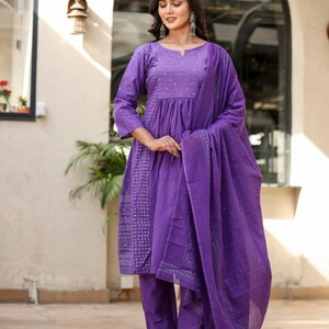 May include: A striking purple salwar kameez outfit. The set features a tunic top with detailed embroidery, paired with straight-leg trousers and a flowing dupatta. The fabric appears lightweight. The model is wearing silver earrings and sandals. The outfit is suitable for warm weather.
