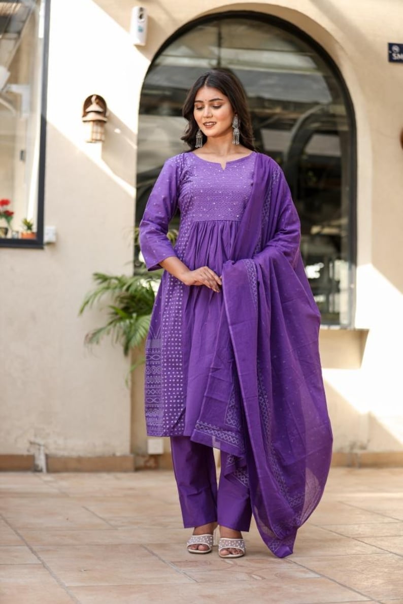 May include: A striking purple salwar kameez outfit. The set features a tunic top with detailed embroidery, paired with straight-leg trousers and a flowing dupatta. The fabric appears lightweight. The model is wearing silver earrings and sandals. The outfit is suitable for warm weather.