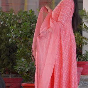 May include: A coral-coloured salwar kameez outfit with a patterned design. The ensemble features a long-sleeved tunic, matching trousers, and a sheer scarf. The fabric has a subtle geometric pattern. The outfit is worn with embellished flats.