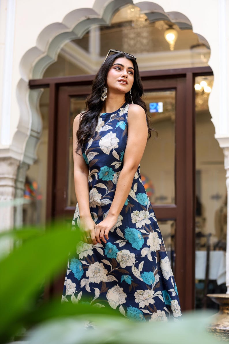 May include: A sleeveless, navy blue floral print dress featuring large white and teal flowers. The dress has a flowing silhouette and is worn by a person standing in front of an arched doorway. Sunglasses rest on the person's head.