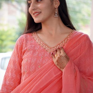 May include: A coral salwar kameez outfit. The top has a V-neck with gold detailing and long sleeves. A matching dupatta is draped over the shoulder. The outfit has a subtle pattern and is worn with silver earrings.