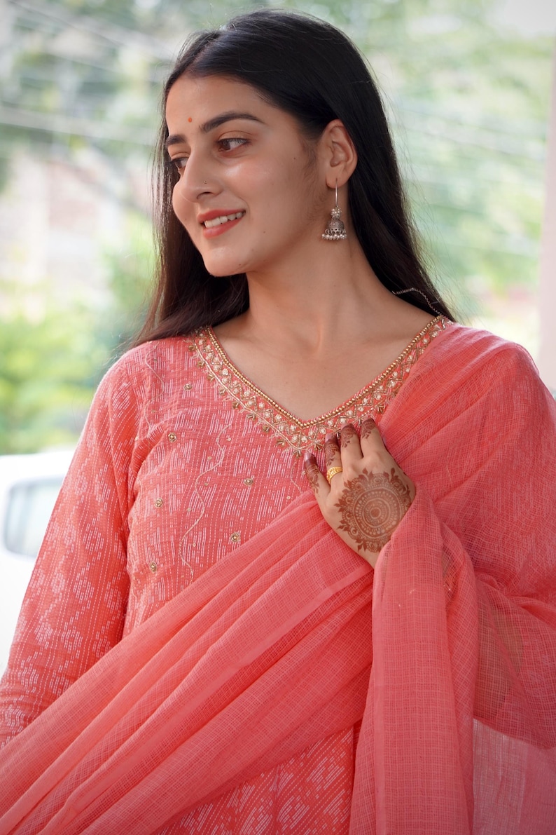 May include: A coral salwar kameez outfit. The top has a V-neck with gold detailing and long sleeves. A matching dupatta is draped over the shoulder. The outfit has a subtle pattern and is worn with silver earrings.