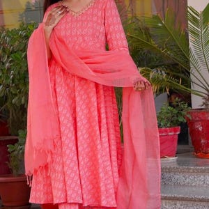 May include: A coral pink Indian salwar kameez outfit. The ensemble includes a long-sleeved tunic with a V-neck, matching trousers, and a sheer scarf. The fabric features a subtle geometric pattern. The outfit is worn with embellished sandals.