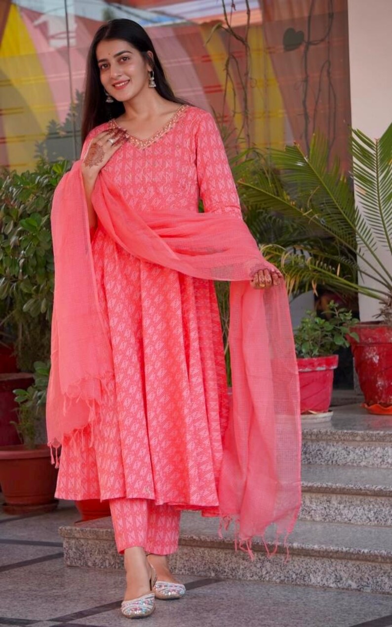 May include: A coral pink Indian salwar kameez outfit. The ensemble includes a long-sleeved tunic with a V-neck, matching trousers, and a sheer scarf. The fabric features a subtle geometric pattern. The outfit is worn with embellished sandals.