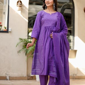 May include: A striking purple salwar kameez outfit. The set features a tunic top with three-quarter sleeves and intricate embroidery, paired with straight-leg trousers and a matching scarf. The lightweight fabric makes it ideal for warmer climates.