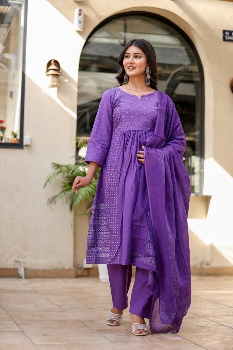 May include: A striking purple salwar kameez outfit. The set features a tunic top with three-quarter sleeves and intricate embroidery, paired with straight-leg trousers and a matching scarf. The lightweight fabric makes it ideal for warmer climates.