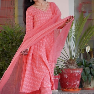 May include: A coral-coloured Indian suit set, comprising a long-sleeved tunic, matching trousers, and a sheer scarf. The tunic has a V-neck with gold detailing and a geometric pattern. The trousers are ankle-length. The outfit is worn with sandals.