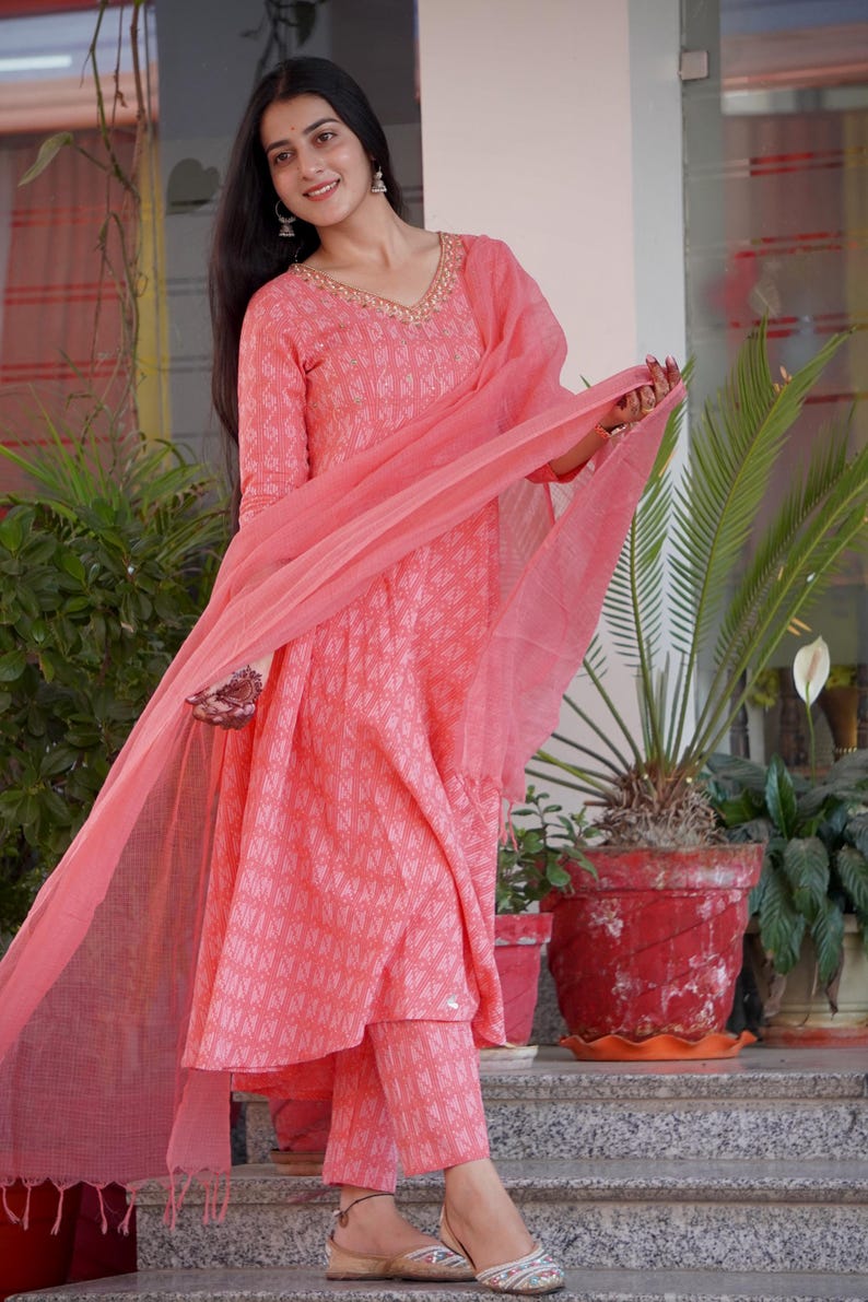 May include: A coral-coloured Indian suit set, comprising a long-sleeved tunic, matching trousers, and a sheer scarf. The tunic has a V-neck with gold detailing and a geometric pattern. The trousers are ankle-length. The outfit is worn with sandals.