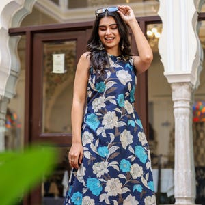 May include: Sleeveless navy blue dress with a floral pattern in shades of white and light blue. The dress is knee-length and has a round neckline. The model is wearing sunglasses and smiling.