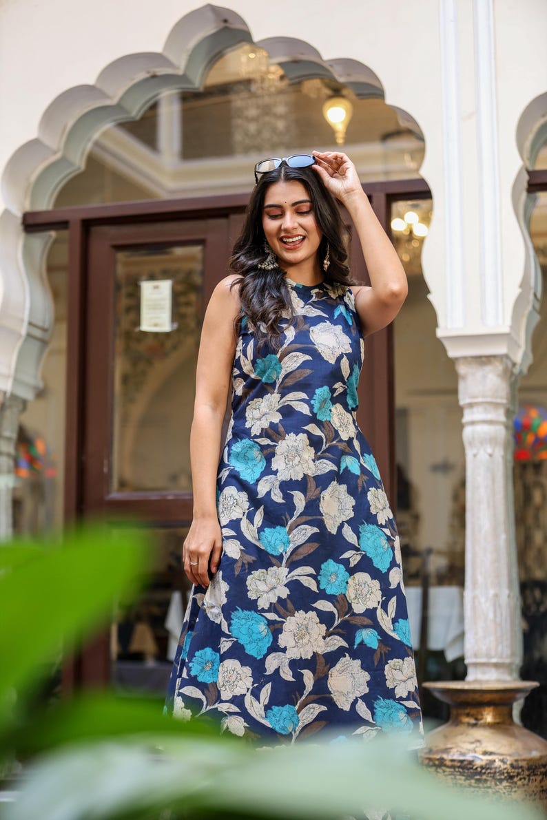 May include: Sleeveless navy blue dress with a floral pattern in shades of white and light blue. The dress is knee-length and has a round neckline. The model is wearing sunglasses and smiling.
