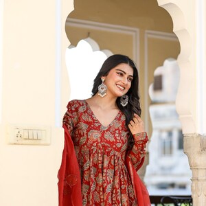 Puede incluir: Un vestido rojo con estampado floral, cuello en V y mangas largas. El vestido se combina con una bufanda roja a juego. La mujer lleva pendientes plateados. El fondo presenta un arco de puerta.