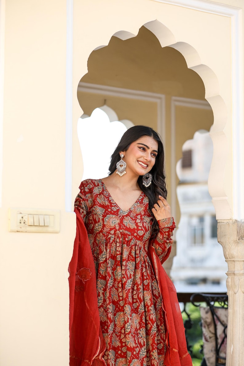 Puede incluir: Un vestido rojo con estampado floral, cuello en V y mangas largas. El vestido se combina con una bufanda roja a juego. La mujer lleva pendientes plateados. El fondo presenta un arco de puerta.