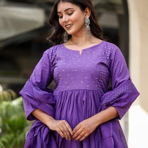 May include: A long, purple dress with a V-neck and long sleeves. The dress has detailed embroidery on the bodice and sleeves. A matching scarf is draped over the dress. The dress is made of a soft fabric.