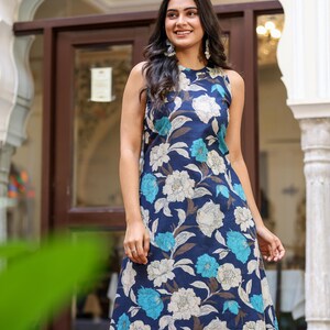May include: Sleeveless navy blue dress with a floral pattern in white, light blue, and brown. The dress has a round neckline and a flowing silhouette. Sunglasses are perched on top of the dress. The dress is suitable for casual or semi-formal occasions.