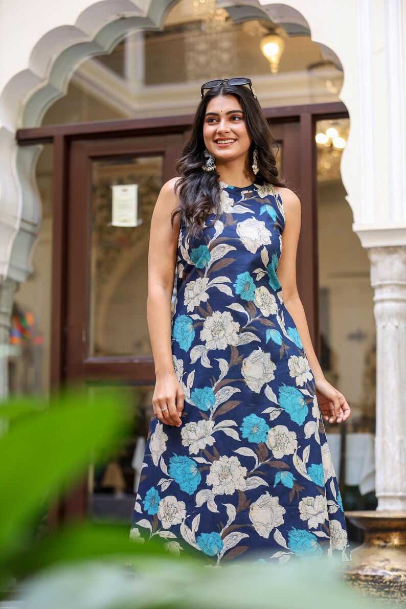 May include: Sleeveless navy blue dress with a floral pattern in white, light blue, and brown. The dress has a round neckline and a flowing silhouette. Sunglasses are perched on top of the dress. The dress is suitable for casual or semi-formal occasions.