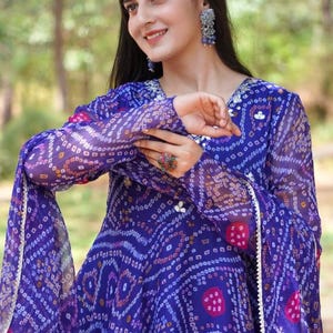 Puede incluir: Un vibrante vestido azul con un patrón detallado de puntos y líneas blancos, rosas y amarillos. El vestido tiene mangas largas y un diseño fluido. El escote está adornado con pequeños detalles blancos. El vestido se usa con aretes y un anillo a juego.