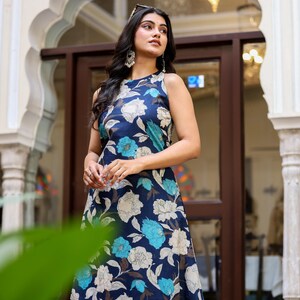 May include: A sleeveless, navy blue floral dress featuring large white and turquoise flowers. The dress has a flowing silhouette and is worn by a person. The background includes architectural details and natural light.