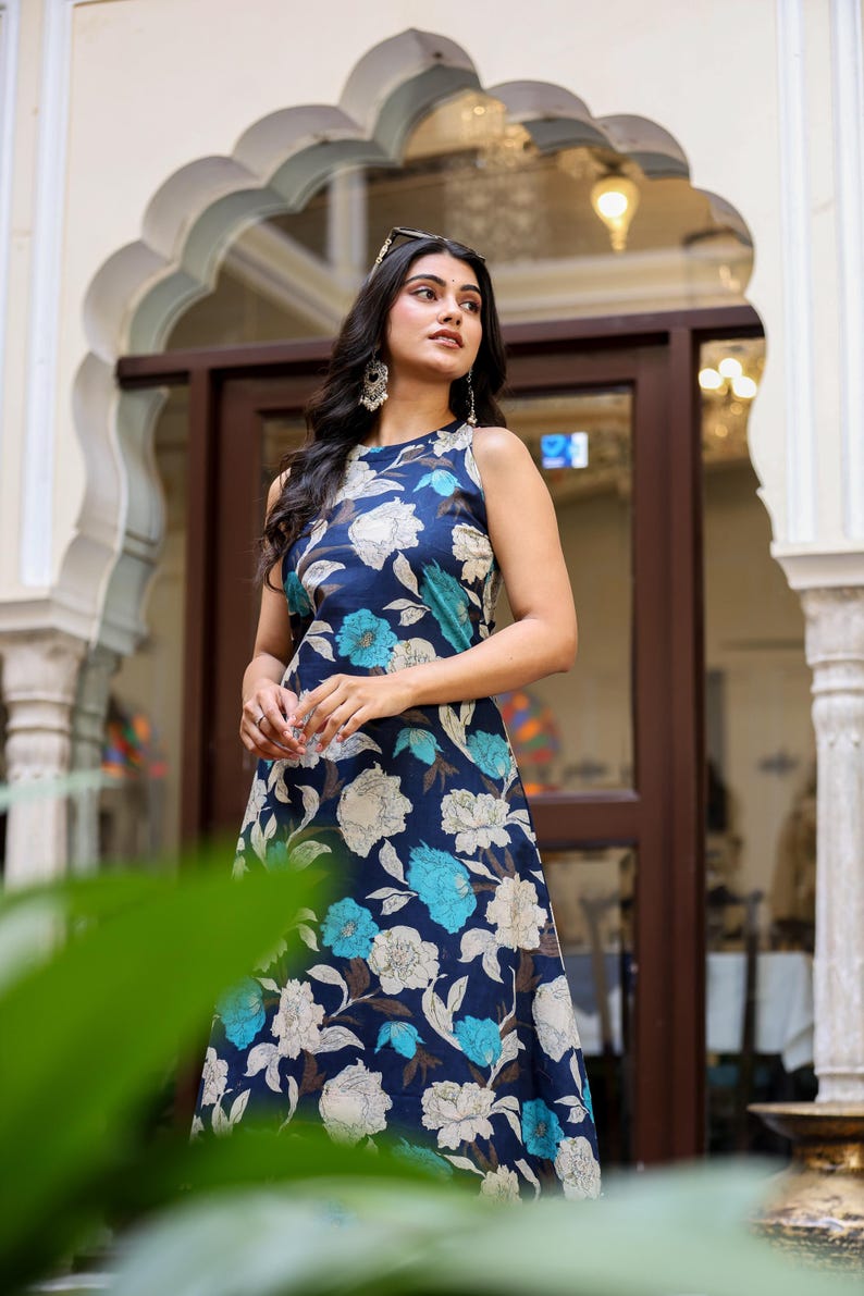 May include: A sleeveless, navy blue floral dress featuring large white and turquoise flowers. The dress has a flowing silhouette and is worn by a person. The background includes architectural details and natural light.