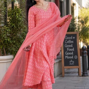May include: A woman in a coral-coloured Indian suit, comprising a long-sleeved tunic, matching trousers, and a sheer scarf. The suit features a geometric pattern and gold detailing around the neckline. The outfit is paired with flat sandals. A chalkboard sign displays "Good Coffee Good Food Good Mood."
