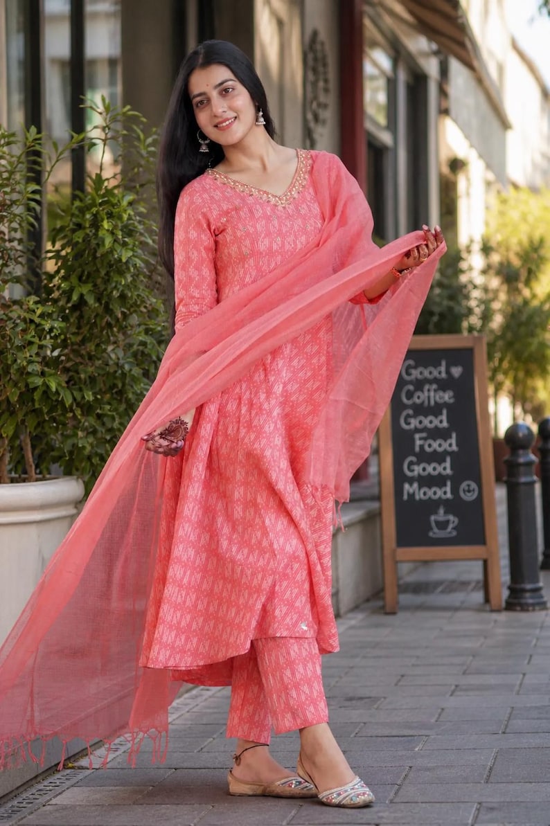 May include: A woman in a coral-coloured Indian suit, comprising a long-sleeved tunic, matching trousers, and a sheer scarf. The suit features a geometric pattern and gold detailing around the neckline. The outfit is paired with flat sandals. A chalkboard sign displays "Good Coffee Good Food Good Mood."