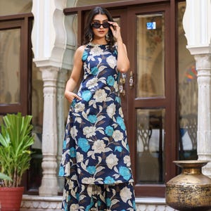 May include: A sleeveless, floor-length dress and matching pants set with a floral pattern in shades of blue, white, and brown. The outfit features a high neckline and a layered skirt. The model is wearing sunglasses and gold heels.