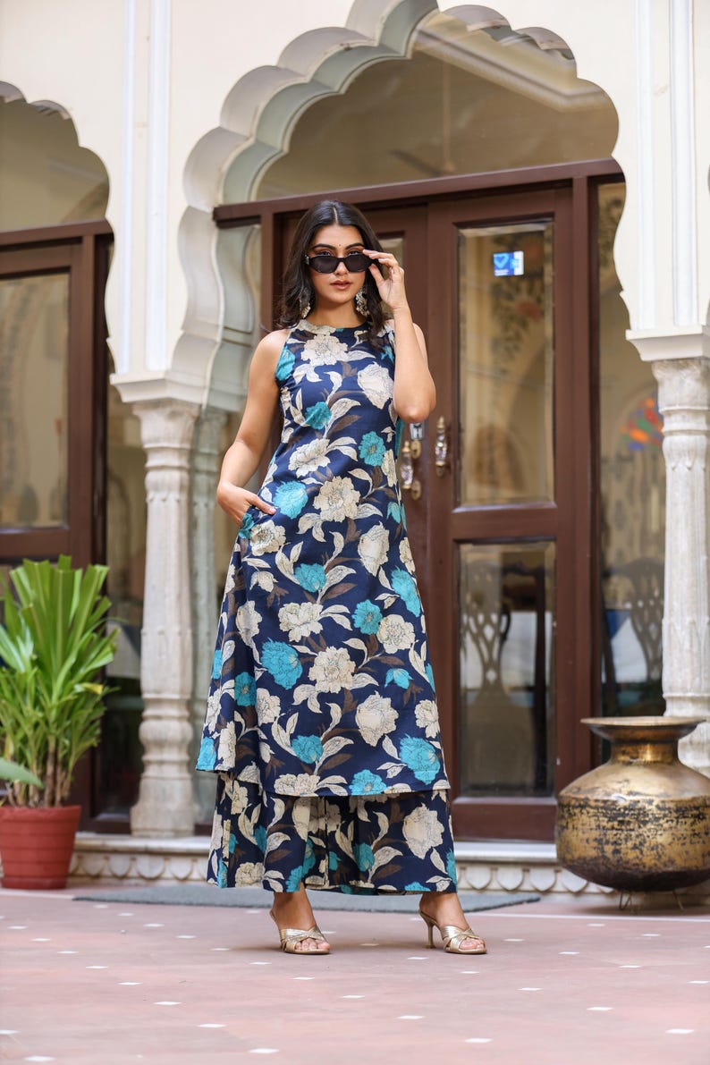 May include: A sleeveless, floor-length dress and matching pants set with a floral pattern in shades of blue, white, and brown. The outfit features a high neckline and a layered skirt. The model is wearing sunglasses and gold heels.