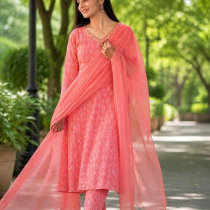May include: A coral-coloured Indian salwar kameez outfit. It features a long-sleeved tunic, matching trousers, and a sheer dupatta. The tunic and trousers have a subtle patterned design, and the neckline is embellished with gold detailing.