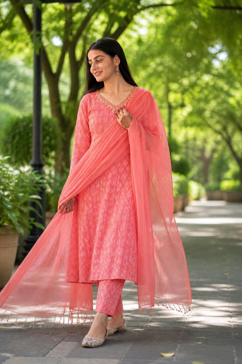 May include: A coral-coloured Indian salwar kameez outfit. It features a long-sleeved tunic, matching trousers, and a sheer dupatta. The tunic and trousers have a subtle patterned design, and the neckline is embellished with gold detailing.