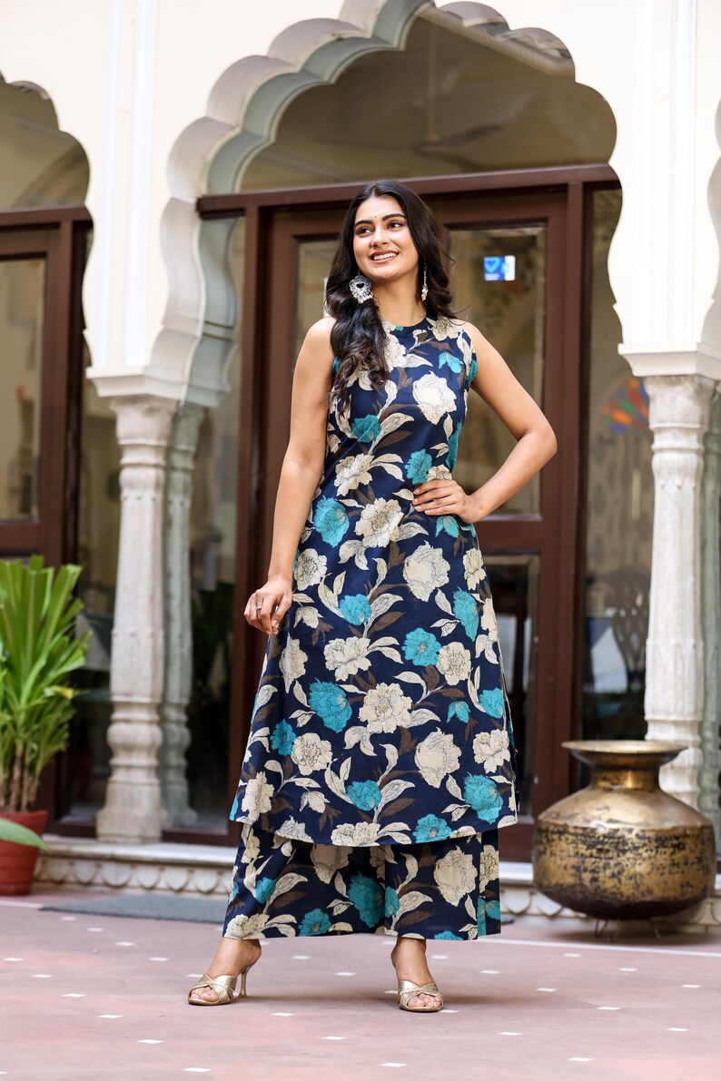 May include: A sleeveless, floral-print outfit in navy blue, light blue, and cream. The ensemble includes a top and matching wide-leg pants. The outfit is worn with gold heels. The background features architectural details.