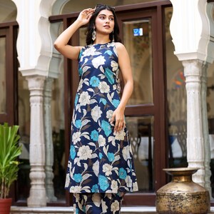 May include: A sleeveless navy blue floral print outfit, including a long tunic and matching pants. The floral pattern features white and light blue flowers. The outfit is worn with gold heels and sunglasses.