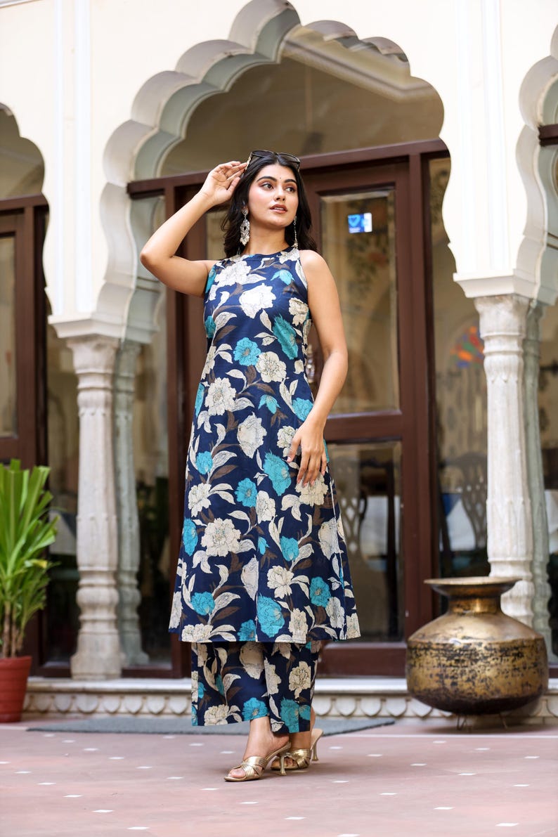 May include: A sleeveless navy blue floral print outfit, including a long tunic and matching pants. The floral pattern features white and light blue flowers. The outfit is worn with gold heels and sunglasses.