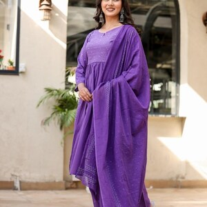 May include: A woman in a striking purple salwar kameez. The outfit comprises a long-sleeved tunic, matching trousers, and a draped scarf. The fabric features a delicate pattern. The woman is wearing silver earrings and sandals.