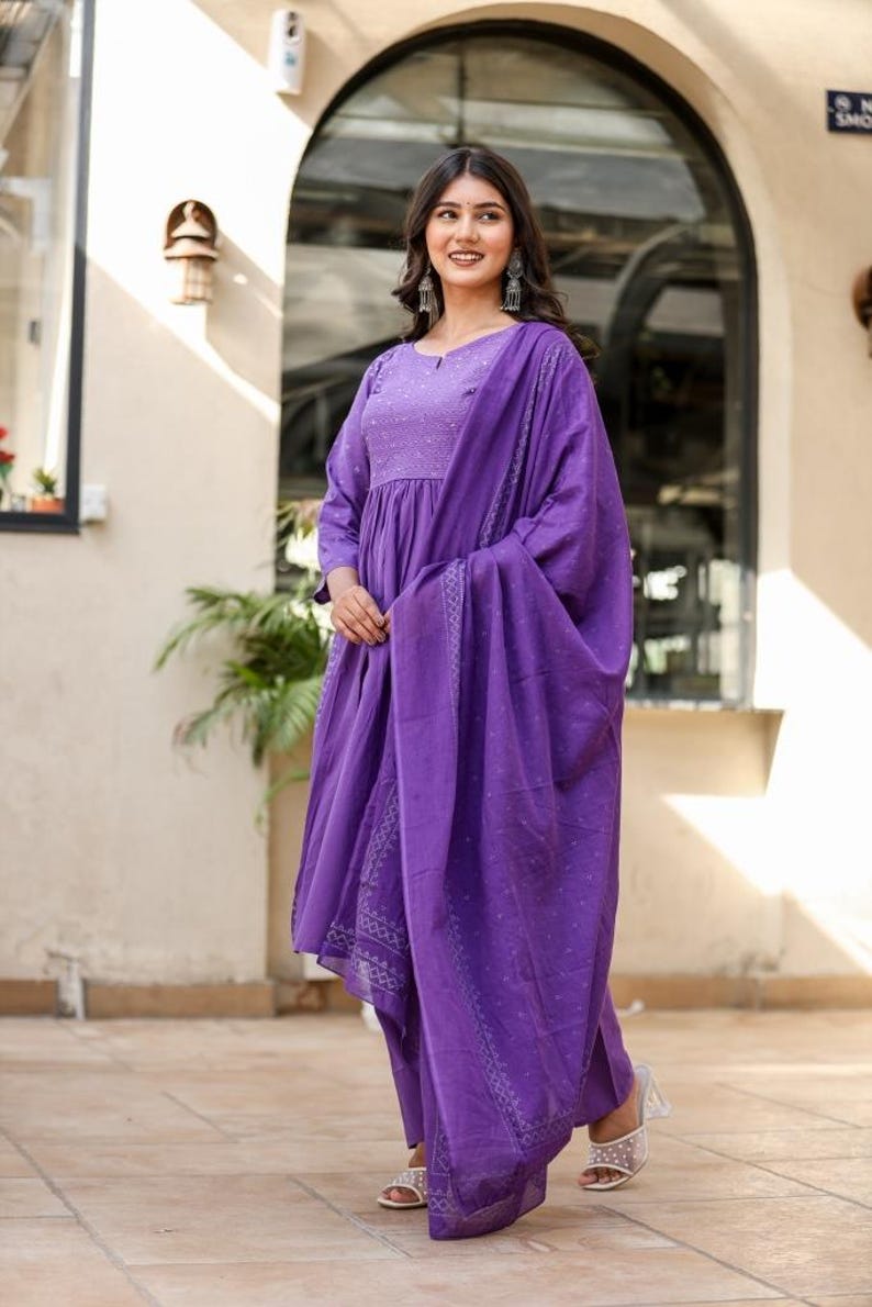 May include: A woman in a striking purple salwar kameez. The outfit comprises a long-sleeved tunic, matching trousers, and a draped scarf. The fabric features a delicate pattern. The woman is wearing silver earrings and sandals.