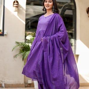 May include: A woman in a striking purple salwar kameez. The outfit comprises a long-sleeved tunic, trousers, and a flowing dupatta. The fabric features a delicate dotted design and decorative borders.