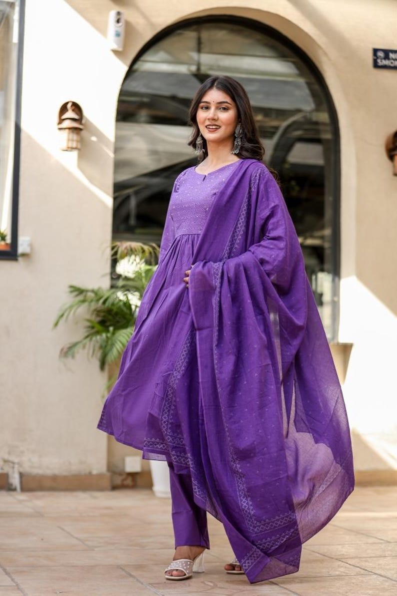 May include: A woman in a striking purple salwar kameez. The outfit comprises a long-sleeved tunic, trousers, and a flowing dupatta. The fabric features a delicate dotted design and decorative borders.