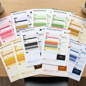 Op de afbeelding: Een verzameling kleurrijke focusplanners, waaronder "Self-Love Daily Planner" en "My Neurodivergent Planner", op een houten tafel. Een kopje koffie en een vetplant zijn ook zichtbaar.