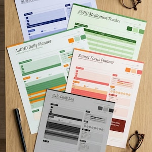 May include: A collection of printed planners, including "ADHD Sleep + Focus Planner," "ADHD Medication Tracker," and "AuDHD Daily Planner." The planners feature color-coded sections and charts. A black pen and two pairs of eyeglasses are also visible on a wooden surface.