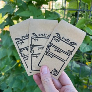 May include: Three kraft paper seed packets with black ink illustrations and text. Each packet has the word "seeds" above a box labeled "Type", "Date", and "Notes". A hand holds the packets in front of a green leafy background.