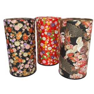 May include: Three cylindrical containers with floral patterns. One is red with white, yellow, and purple flowers. Another is black with orange, pink, and white flowers. The third is black with a crane and peony design.