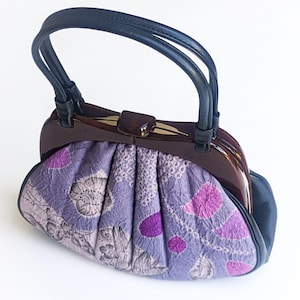 May include: A vintage handbag with a dark brown wooden frame and a black handle. The bag features a pleated, patterned fabric in shades of lavender and purple, with floral and dot designs. The clasp is a dark brown metal.