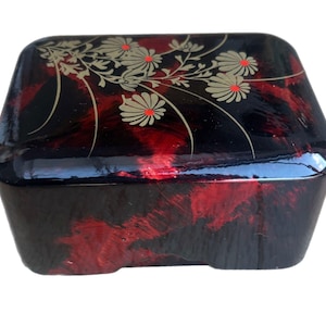 May include: A hexagonal lacquered box with a glossy finish. The box has a black and red marbled pattern. The lid is decorated with a gold floral design with red accents. The box has a low profile and a slightly curved shape.