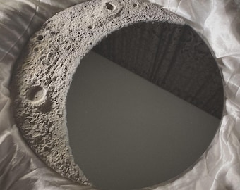 May include: A circular wall mirror featuring a textured, gray, cratered lunar surface along one side, mimicking the appearance of the moon. The remaining portion of the mirror is a smooth, reflective glass surface that creates a crescent moon shape.