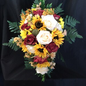May include: A cascading bridal bouquet featuring white roses, burgundy roses, and sunflowers. The bouquet is accented with greenery and small, light brown flowers.