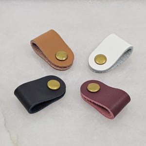 KALLACALLA leather cable organizer clips for charging cords and earphones, made with brass snap closure, shown in black, brown, white, and wine red. Cord management accessories for desk, travel, and everyday carry.