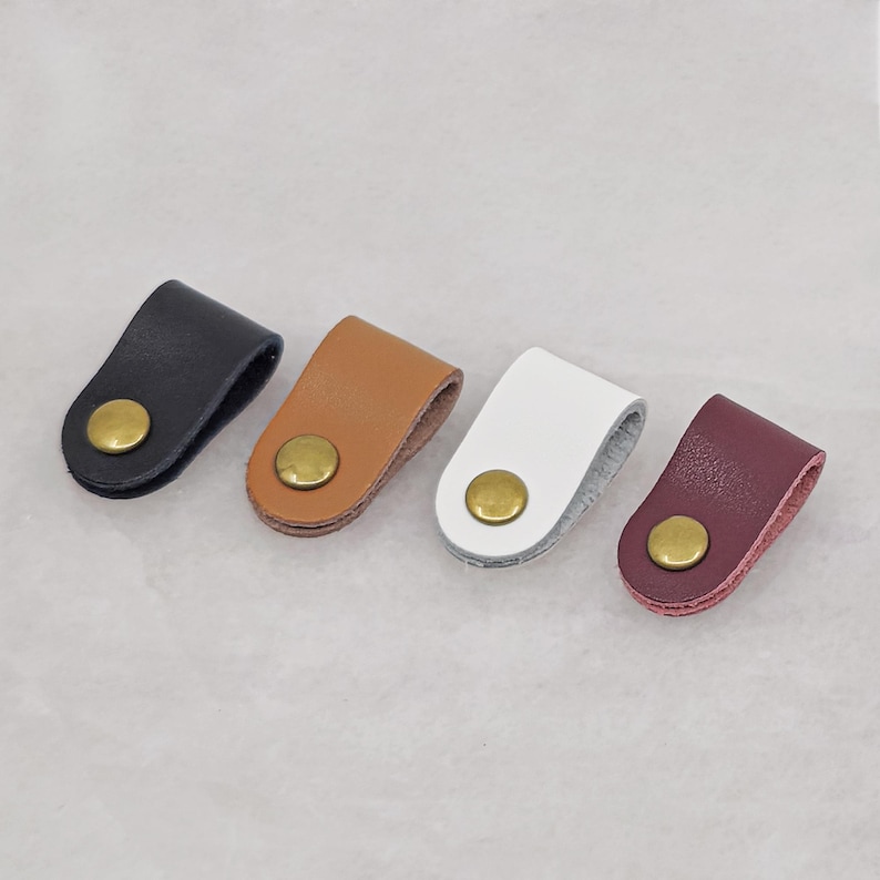 KALLACALLA leather cord organizer clips with brass button snaps, shown in black, tan, white, and wine red. Cable management holder for phone chargers, earbuds, and desktop organization.