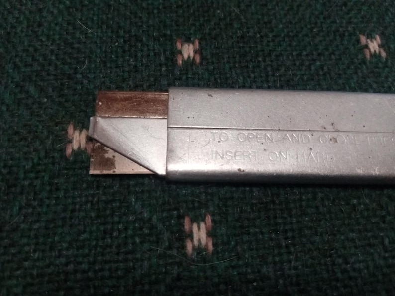 Op de afbeelding: Een zilveren hobbymes met een gedeeltelijk blootliggend mes. Het mes heeft een bruine, roestige uitstraling. De metalen behuizing van het mes heeft de tekst "TO OPEN AND... INSERT ON HARD". Het mes ligt op een donkergroen, getextureerd oppervlak.