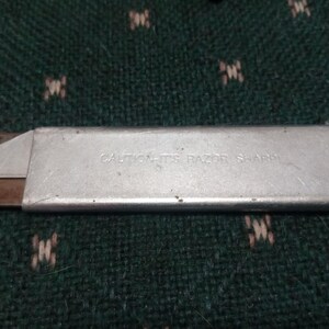 Op de afbeelding: Een zilveren hobbymes met een zichtbaar mes en de tekst "CAUTION-ITS RAZOR SHARP". Het mes ligt op een groen oppervlak met een patroon. Het uiteinde van het mes heeft het woord "MEXICO".